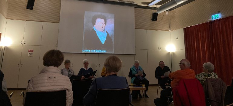 im Hintergrund Videobild Ludwig van Beethoven, Vorleseclub: 5 Vorleser:innen sitzen auf Stühlen und lesen, im Vordergrund Zuschauern auf Stühlen hören zu