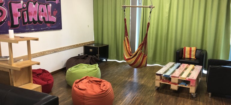 Bildbeschreibung: ein gemütlicher Raum mit Paletten-Tisch und zwei schwarzen Sesseln ist zu sehen. Außerdem gibt es eine Gruppe mit Sitzkissen in verschiedenen Farben und eine Sitzschaukel in herbstlichen Farben. Der grüne Vorhang rundet diesen Raum im gemütlichen Ambiente ab.