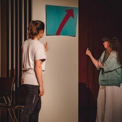 Zwei Frauen stehen mit erhobenem Mittelfinger vor einem blauen Plakat mit rotem Pfeil.