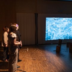 Ein Videofilm mit tosendem Wasser läuft in der Mitte, eine Gruppe von Menschen schaut zu.