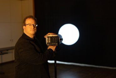Michael Lüdecke schaut in die Kamera und hält einen Schweinwerfer, ein Lichtkegel auf dem Vorhang ist zu sehen