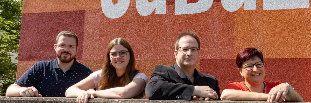 Bildbeschreibung: Zwei Frauen und zwei Männer stehen hinter einer Mauer und beugen sich vor, lächeln in die Kamera. Hinter ihnen ist das Logo des JuBüZ zu sehen.