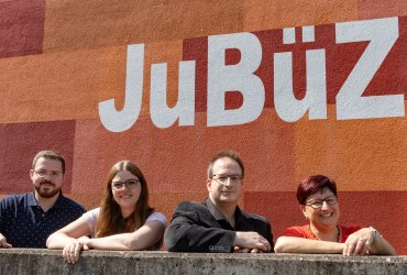 Bildbeschreibung: Zwei Frauen und zwei Männer stehen hinter einer Mauer und beugen sich vor, lächeln in die Kamera. Hinter ihnen ist das Logo des JuBüZ zu sehen.